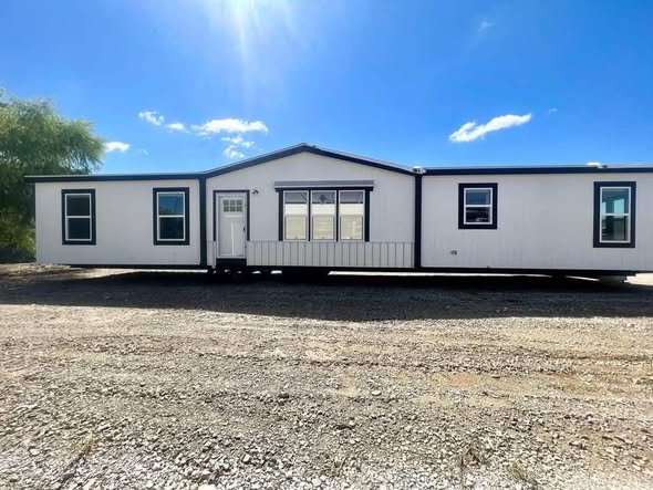 A modern, white manufactured home with black trim, large windows, and a central entryway sits under a bright blue sky with scattered clouds. The scene feels spacious and sunny.