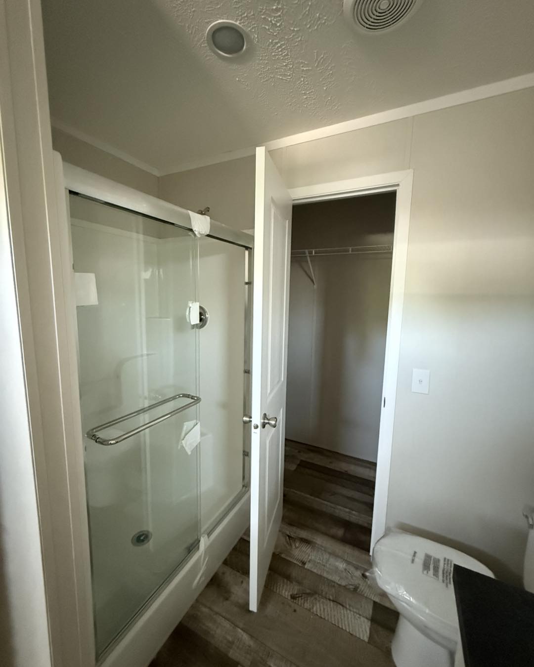 A bathroom with a glass shower door partially open, a closed white closet door, wooden floor, and a toilet with a plastic-wrapped seat.