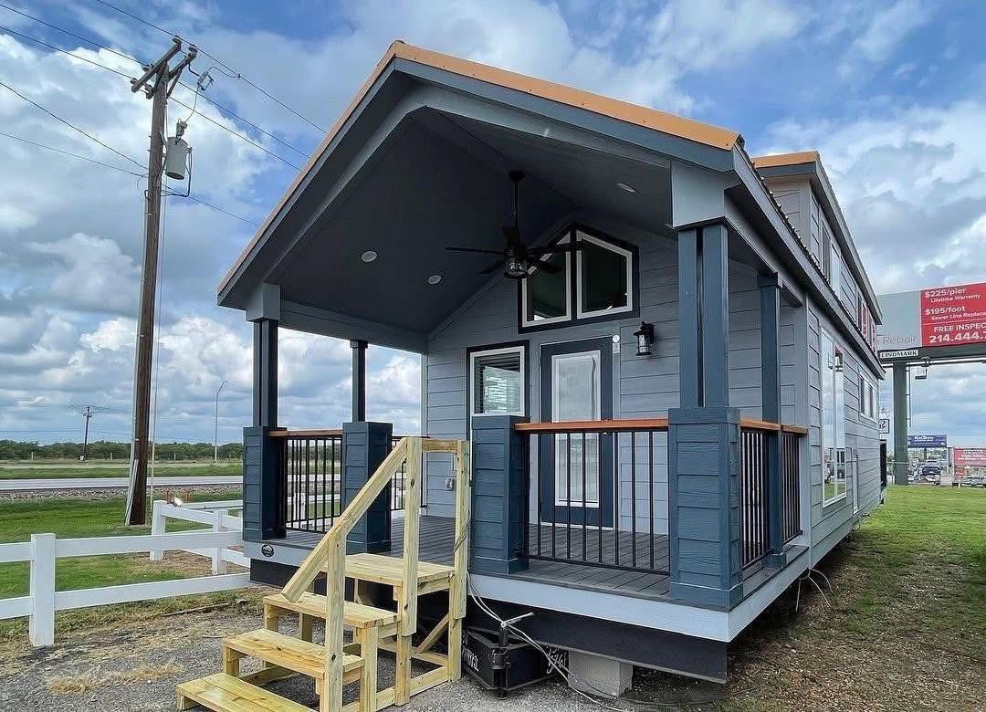 Small blue house with a porch, wood steps, and railing under a cloudy sky. Features a ceiling fan and large windows, set near a road.