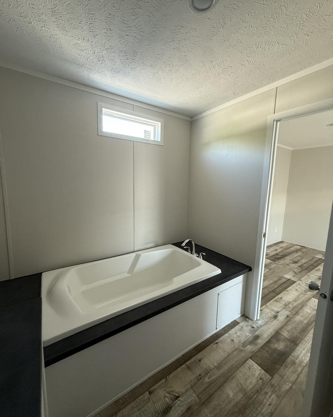 Small bathroom featuring a modern white bathtub with a dark countertop. Sunlight enters through a high window, illuminating the wood-patterned floor.