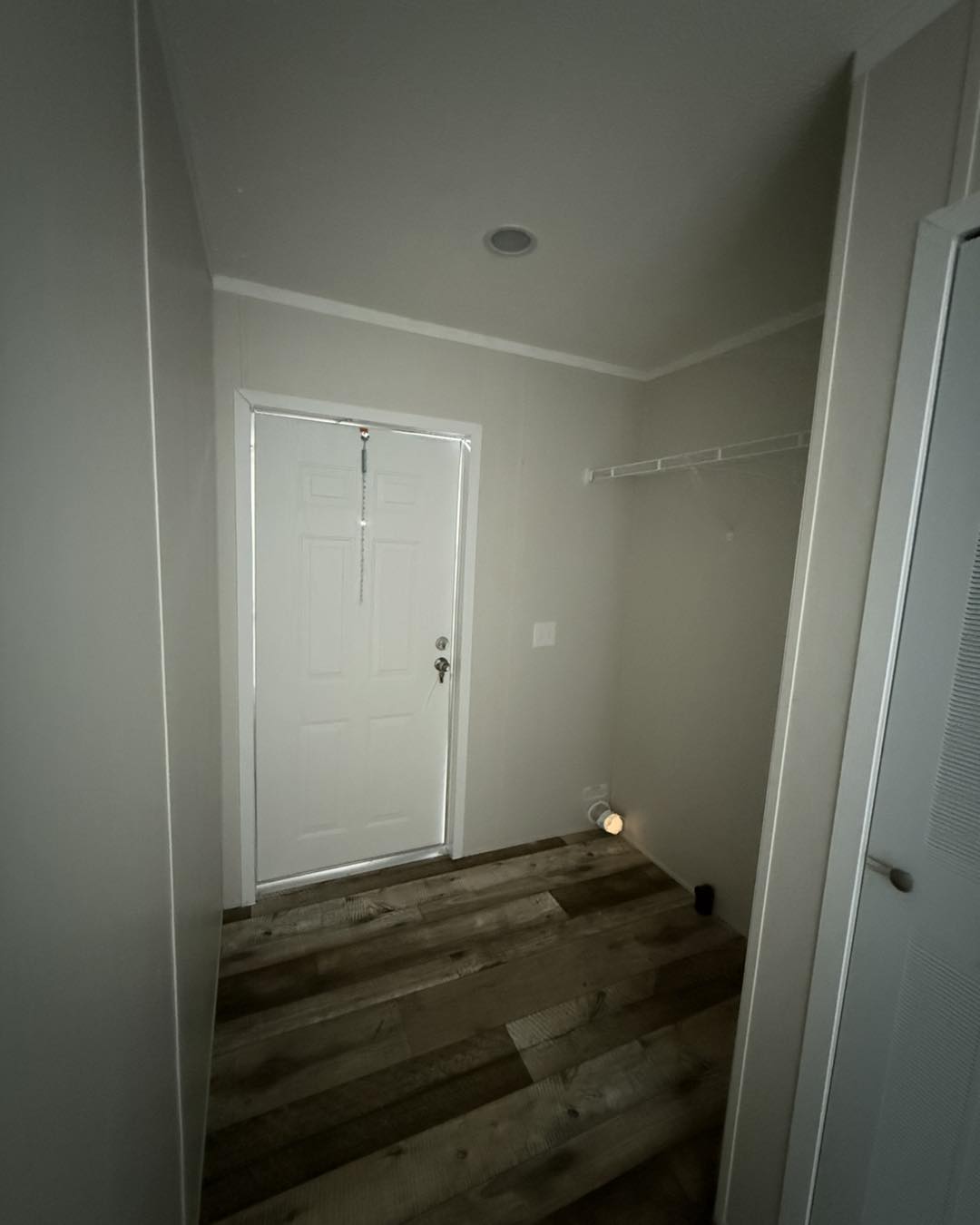 Narrow hallway with a white door in the background, wooden flooring, and beige walls. An empty white shelf is on the right, creating a minimalist feel.