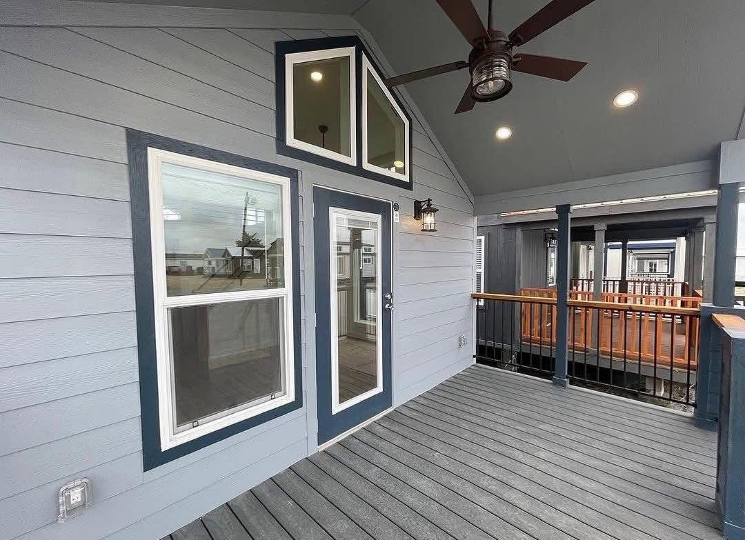 Covered porch with light blue siding, two large windows, and a glass door framed in dark blue. Ceiling fan and lights above; view of adjacent buildings.