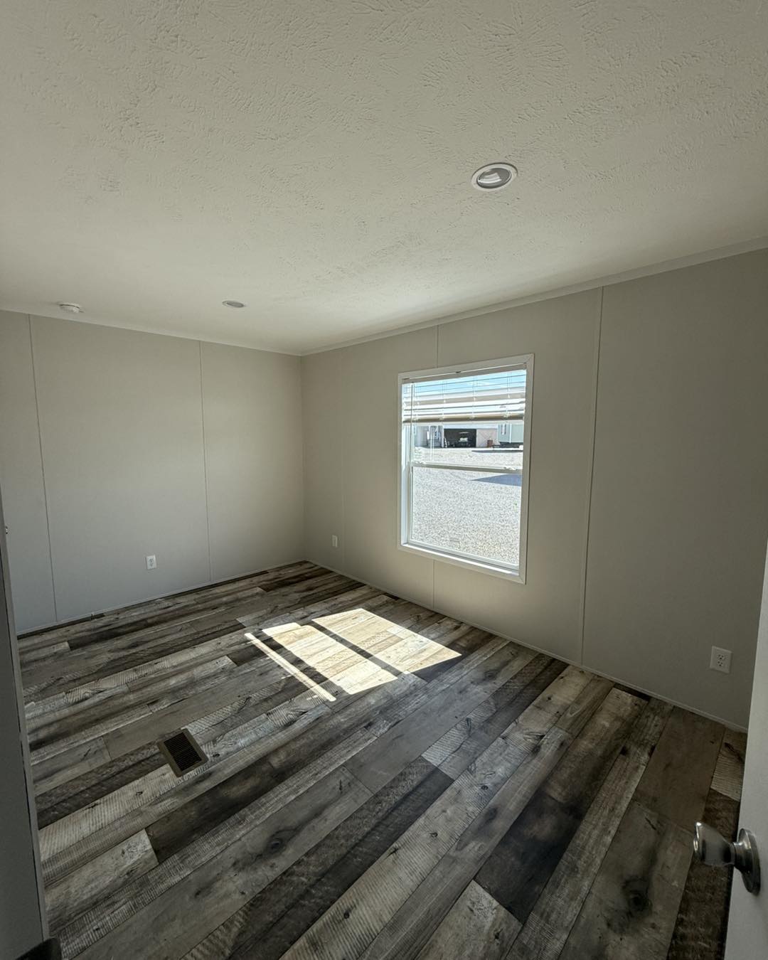 An empty room with light gray walls and a window, featuring rustic wooden flooring. Sunlight casts a pattern on the floor, creating a calm ambiance.
