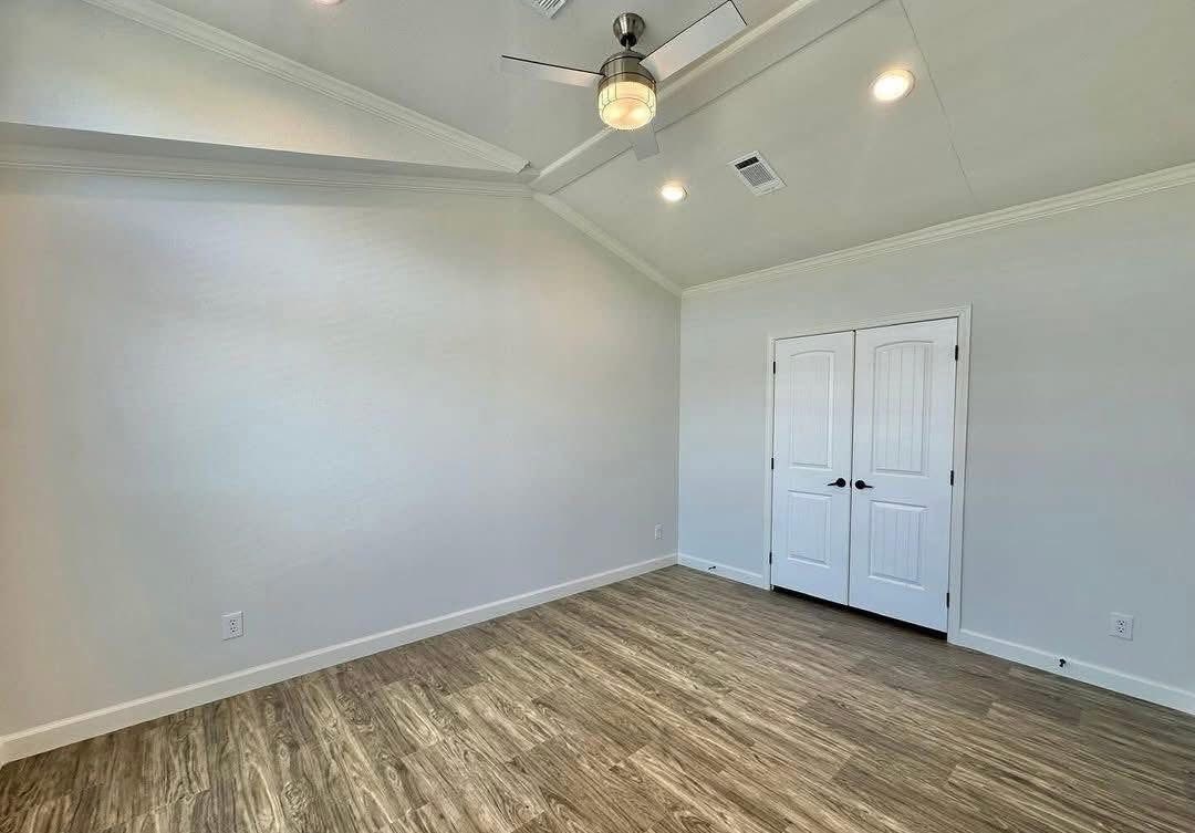 An empty room with light gray walls, wood laminate flooring, and a vaulted ceiling. A ceiling fan with a light is on, and there's a white double door.