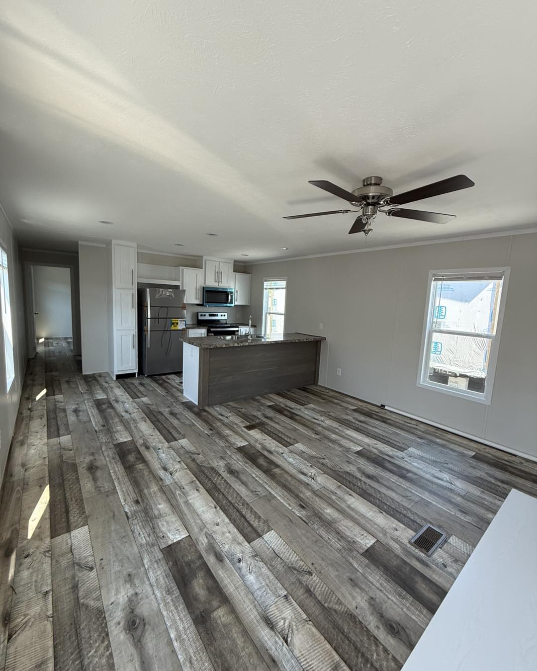 Modern kitchen with white cabinets and stainless steel appliances, light streaming in through windows, wood-patterned flooring, and ceiling fan.