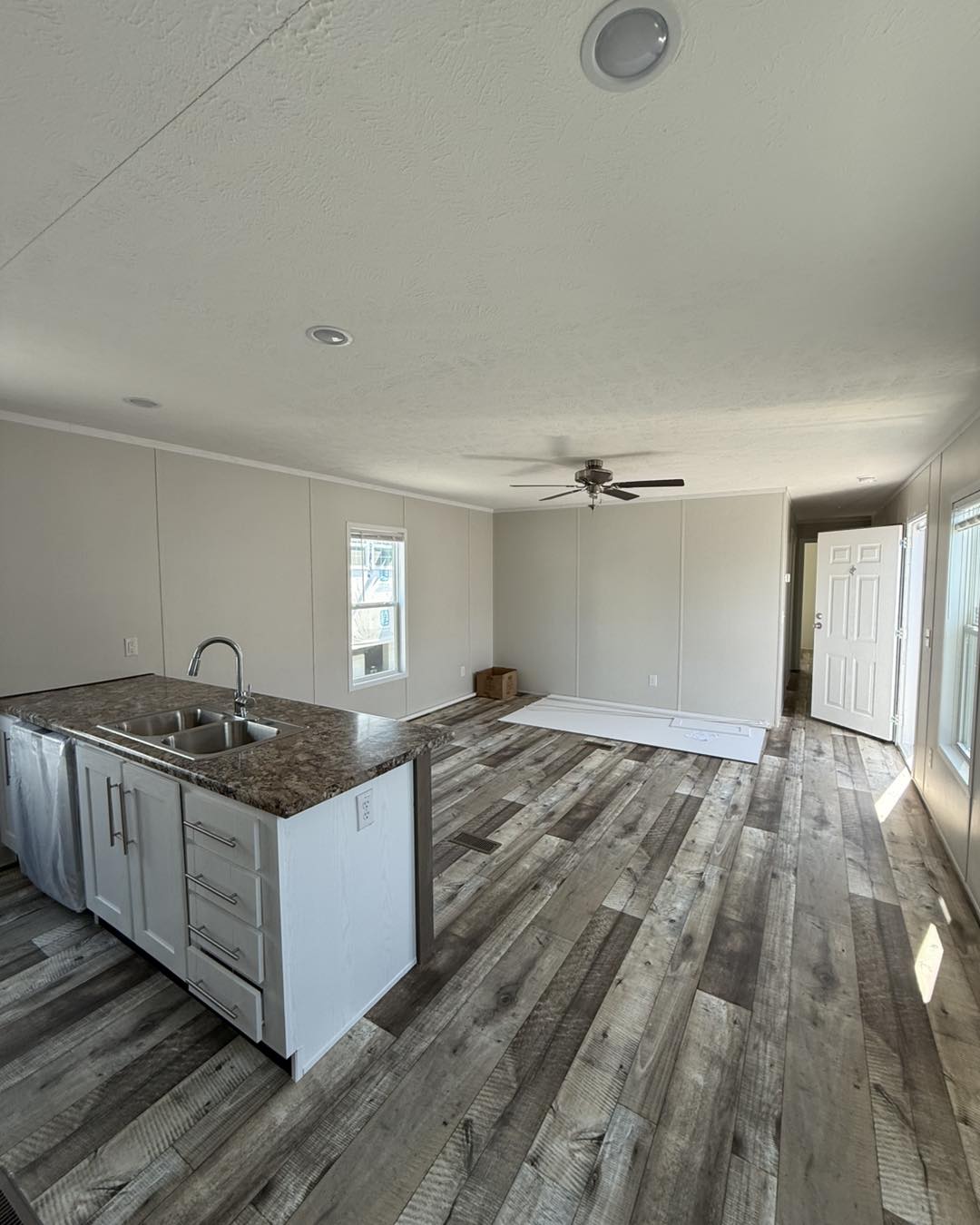 Open, unfurnished room with light wood flooring, white walls, and natural light. Features a kitchen island with a sink and a ceiling fan.