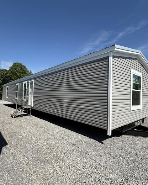 A light gray modular home sits on a gravel lot under a clear blue sky. The home has several white-framed windows and a small set of steps leading to a door.