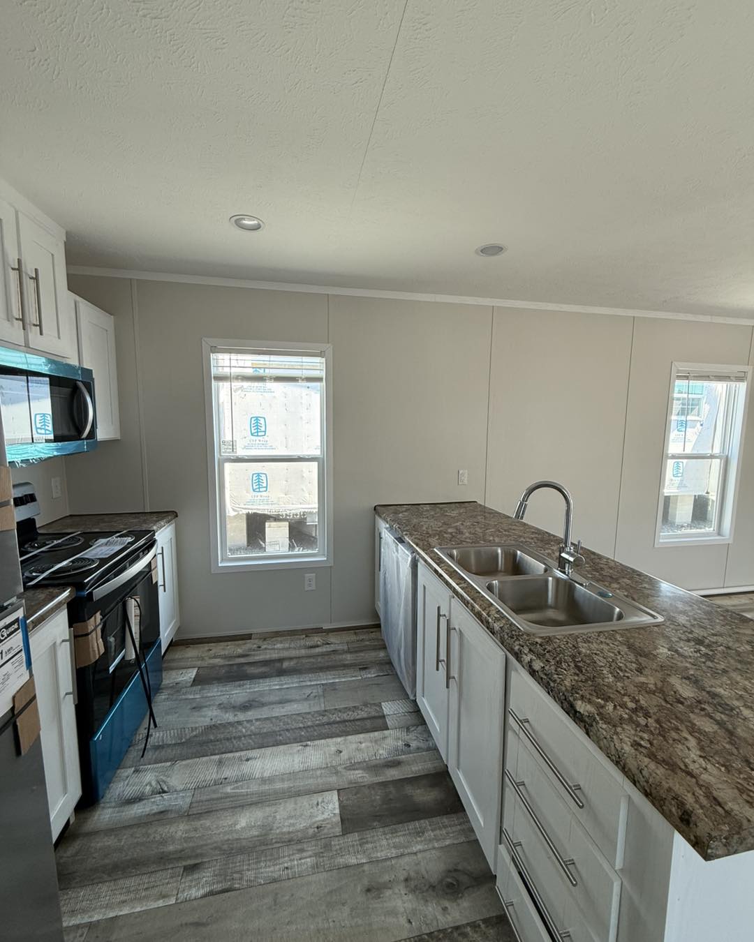 Modern kitchen with light wood flooring, white cabinets, and marble-patterned countertops. A double sink with a silver faucet is visible. Bright natural light from two windows enhances the spacious feel.