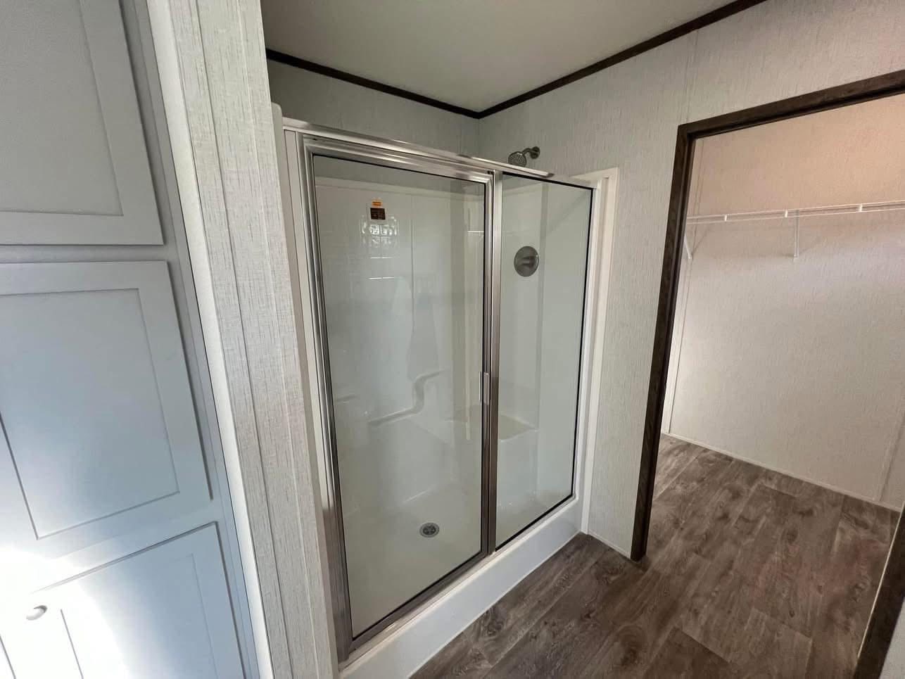 Modern bathroom with a glass shower enclosure, chrome fixtures, and light-colored walls. Adjacent is a walk-in closet with wooden flooring.