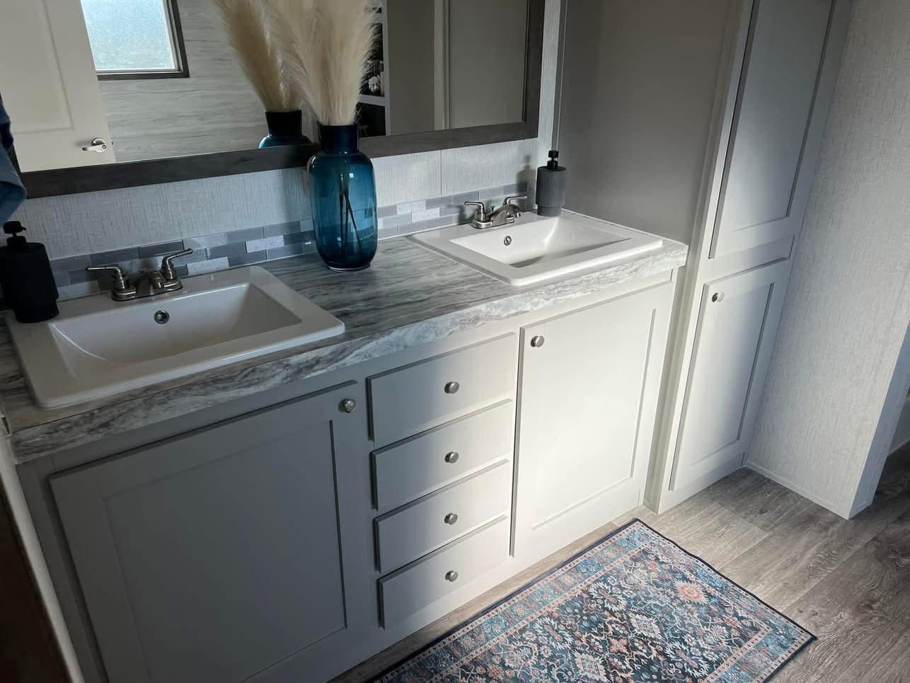 A modern bathroom features a double vanity with gray cabinets, a marble countertop, and two sinks. A blue vase with pampas grass adds an elegant touch, along with a decorative rug on wood flooring.