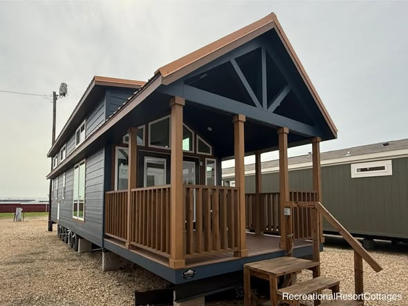 A modern tiny house with a dark blue exterior and brown accents, featuring a small front porch with wooden railings, set on a gravel lot under a cloudy sky.