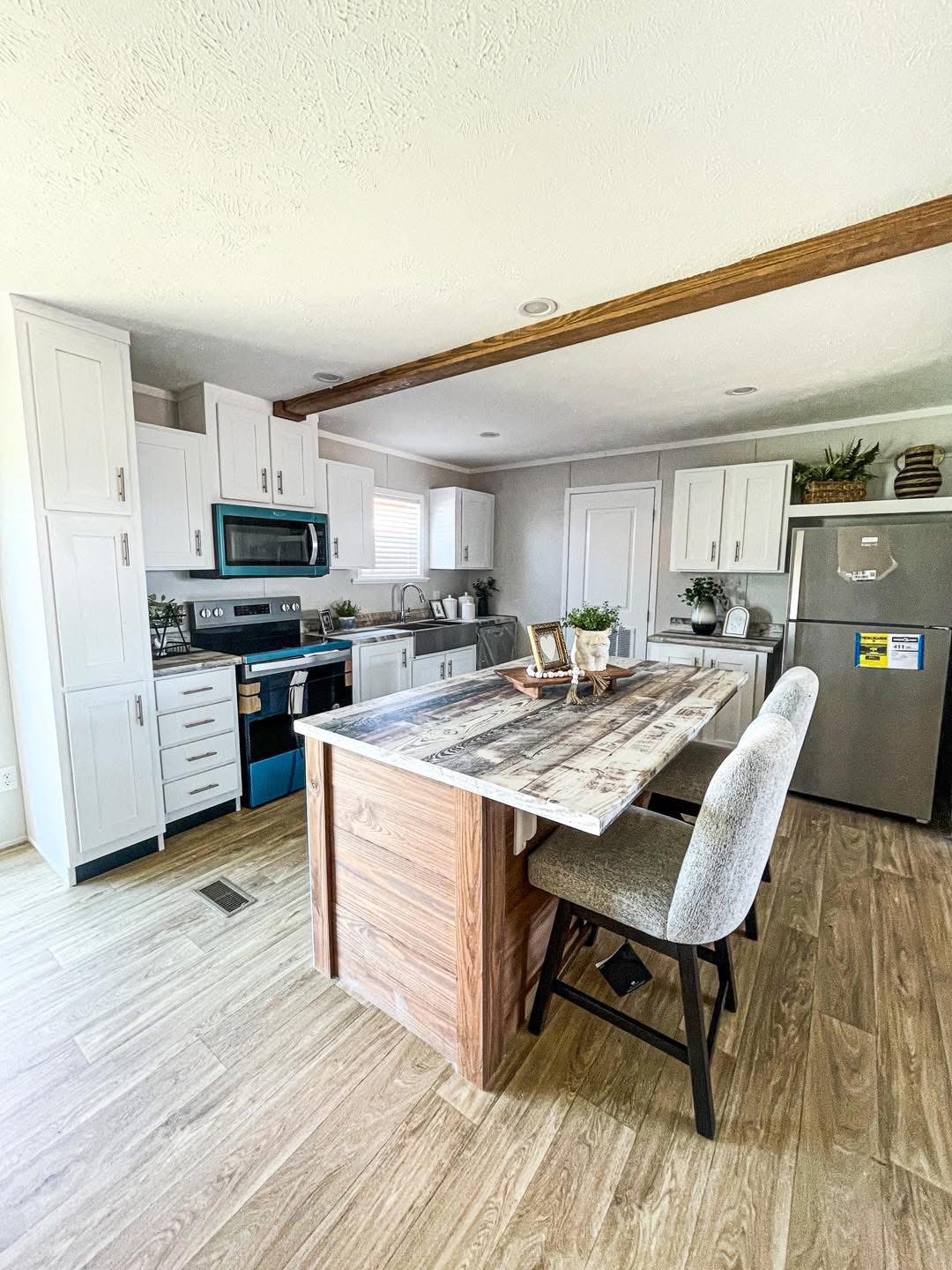 Modern kitchen with light wood flooring, white cabinets, and stainless steel appliances. A rustic island with chairs centers the space, evoking a cozy vibe.
