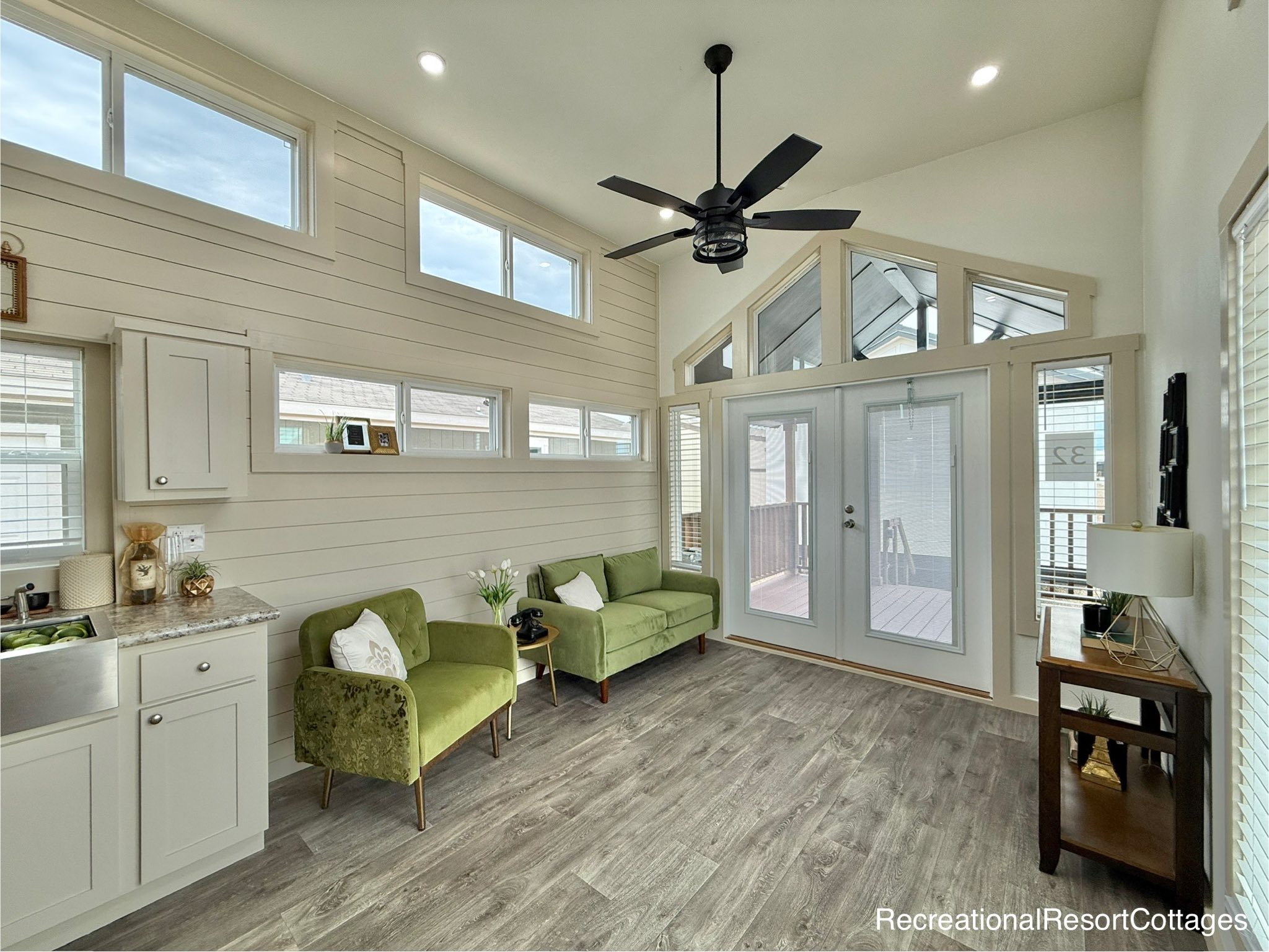 Bright, modern living room with high ceilings, white shiplap walls, and large windows. Features green sofas, overhead fan, and glass doors.