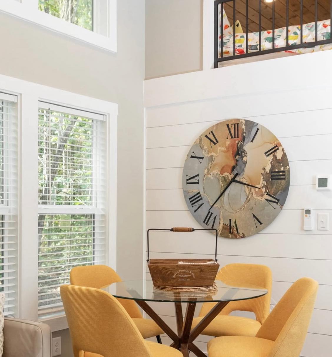 A modern dining area with four mustard-yellow chairs around a round glass table. A large, artistic wall clock shows Roman numerals. Soft natural light streams in through the window blinds, creating a cozy ambiance.