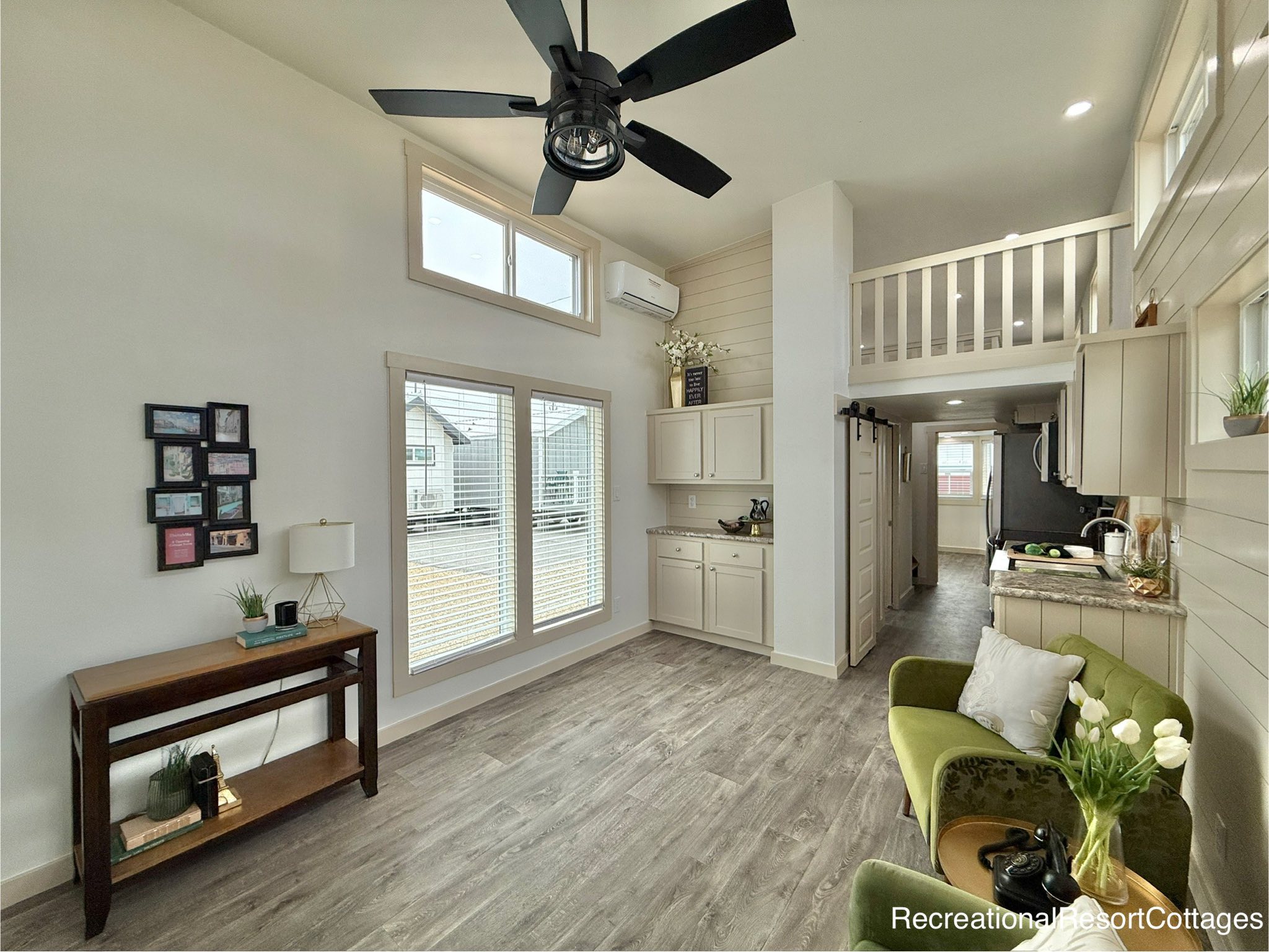 Modern tiny home interior with high ceilings, featuring a ceiling fan, large windows, loft area, and airy decor. A green armchair sits near a cozy kitchen.