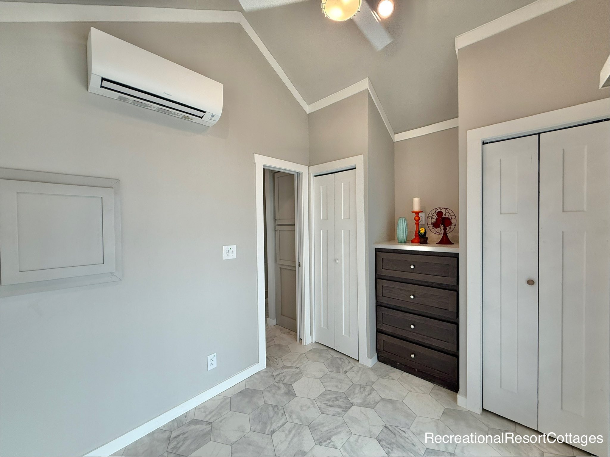 Compact, modern room with beige walls and geometric tiled floor. Features a wall-mounted air conditioner, dark dresser with décor, white closet doors, and recessed lighting. Calm and minimalist.