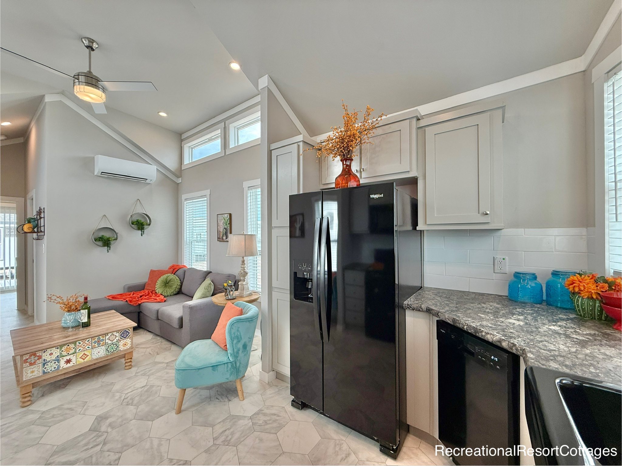 Bright, modern living room and kitchen with vaulted ceilings. A cozy gray sofa with colorful cushions sits by large windows. A black fridge and patterned coffee table add contrast, while vibrant decor gives a welcoming feel.