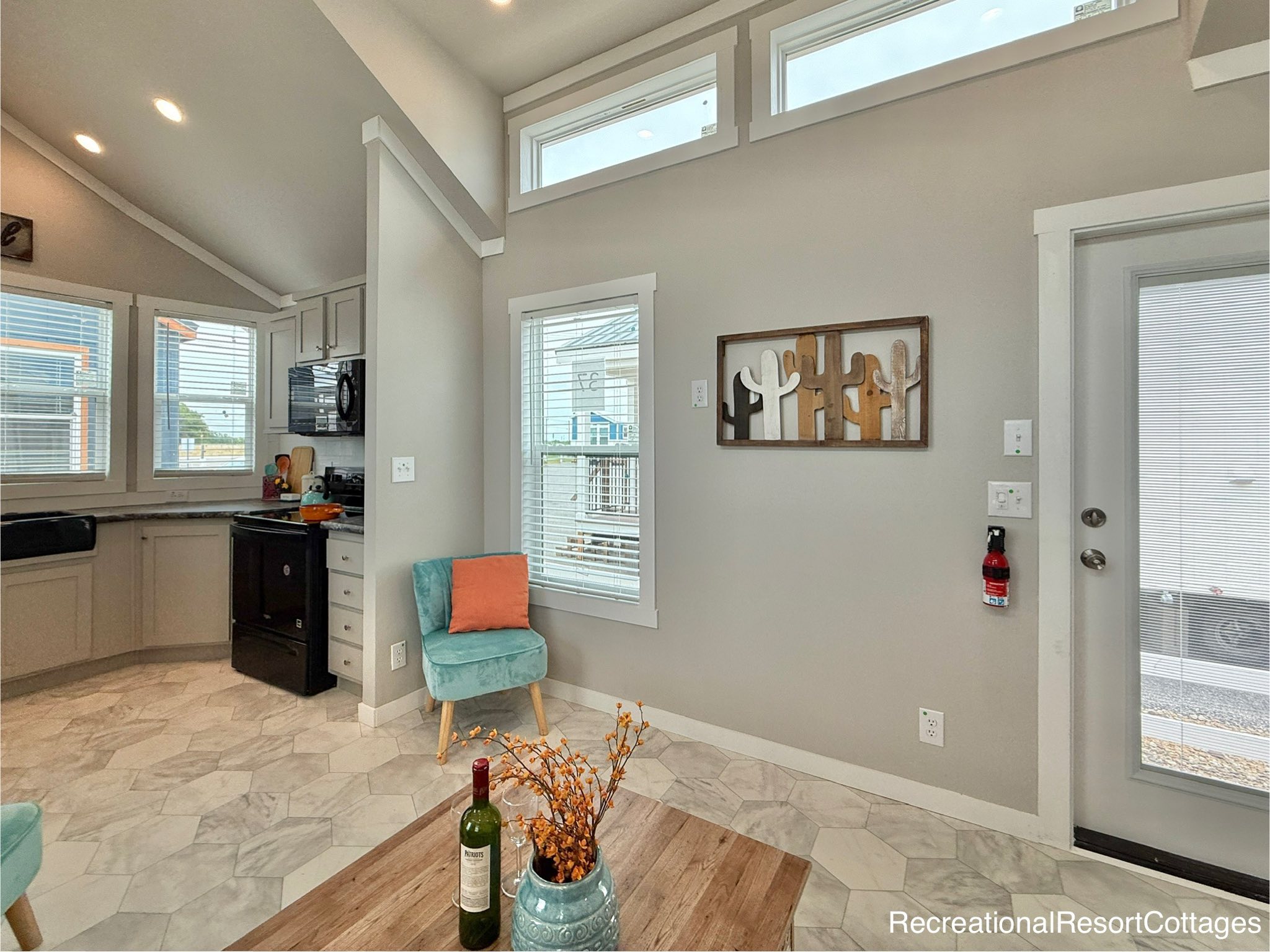 Bright, modern living area with light gray walls and hexagonal tile floor. A teal chair and cactus wall art add charm. Tall windows let in natural light.