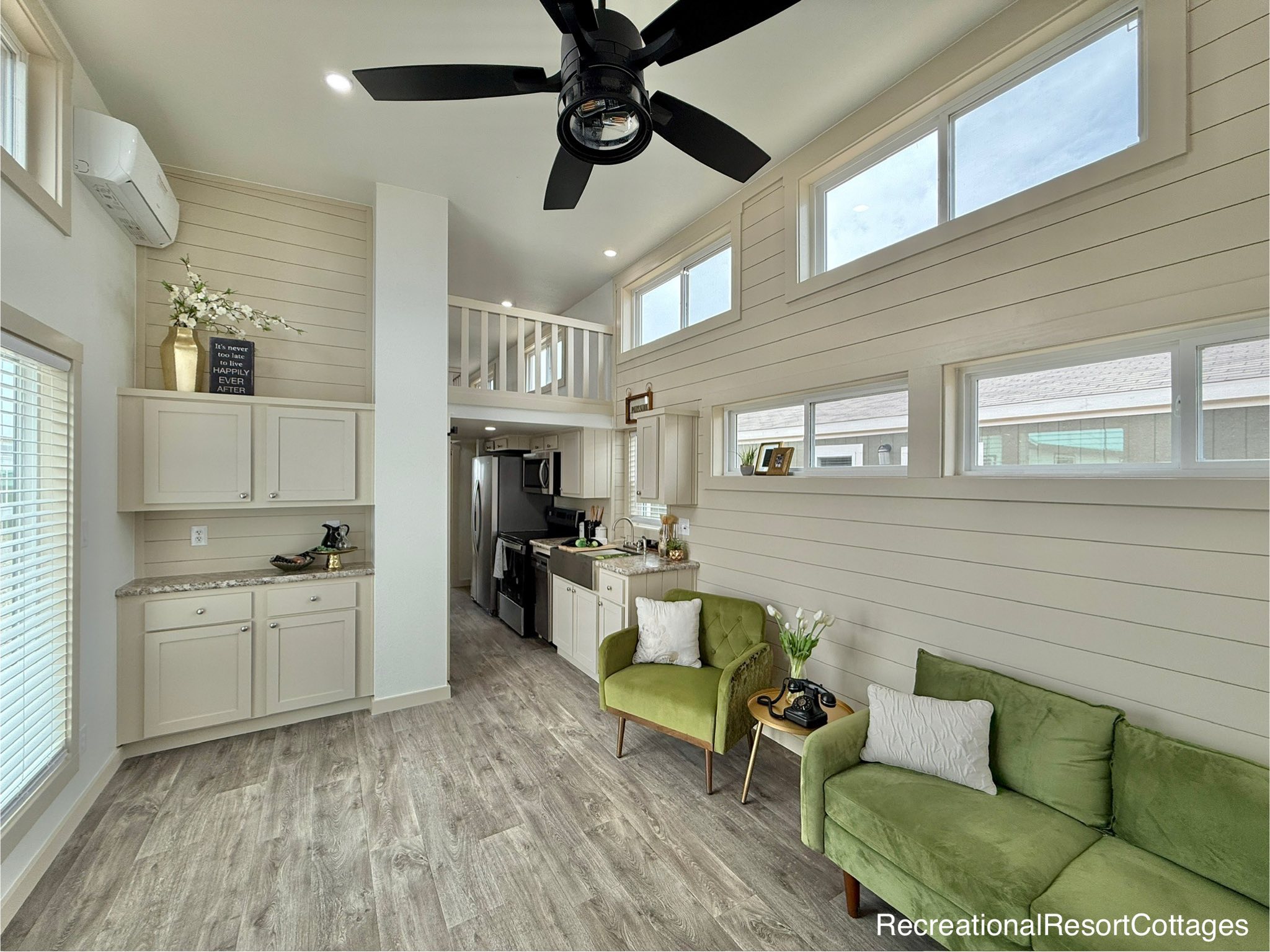 Modern tiny home interior with high ceiling and loft. Features light wood flooring, elegant green chairs, a sleek kitchen, and ample natural light.