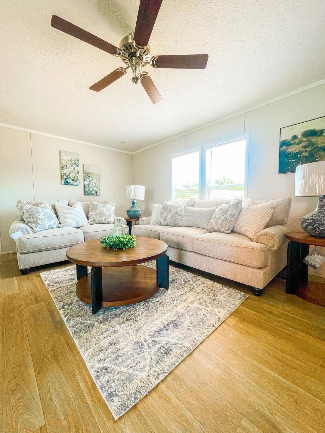 A cozy living room with light beige sofas adorned with patterned cushions, a round wooden coffee table with a plant centerpiece, a textured rug, and elegant decor under a ceiling fan.