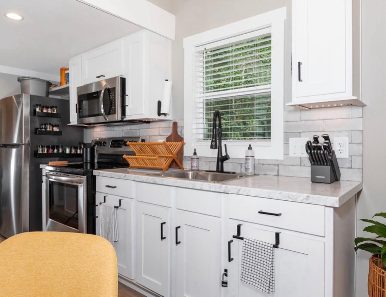 Modern kitchen with white cabinets, marble countertops, and stainless steel appliances. A wooden dish rack is beside the sink under a window.