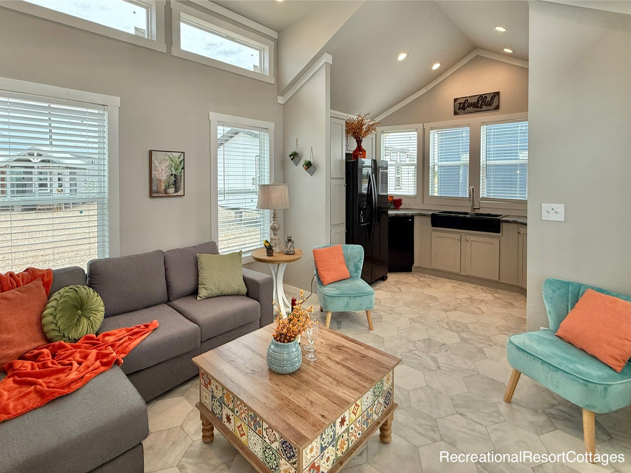 Airy living room with vaulted ceiling, gray sofa with green and orange cushions, colorful coffee table, two teal chairs, and a modern kitchen.