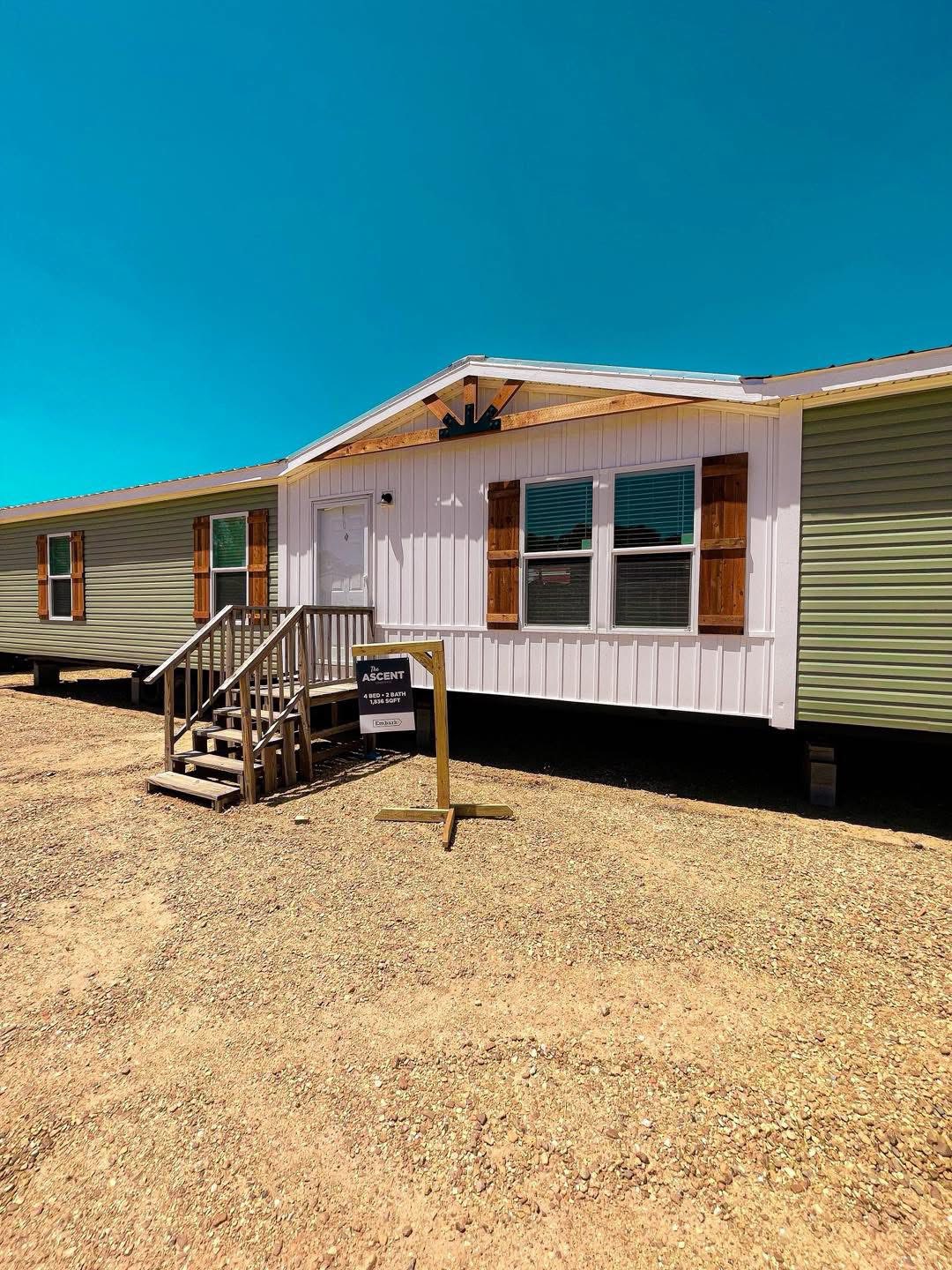 A single-story manufactured home with light green siding and wood accents is set against a clear blue sky. A small sign is in front by the wooden steps.