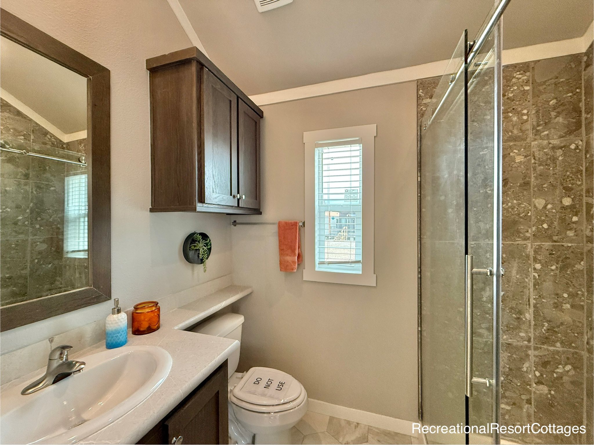 Compact, modern bathroom with natural tones. Features a white sink, dark wood cabinets, glass shower, window with blinds, and orange towel. Cozy ambiance.