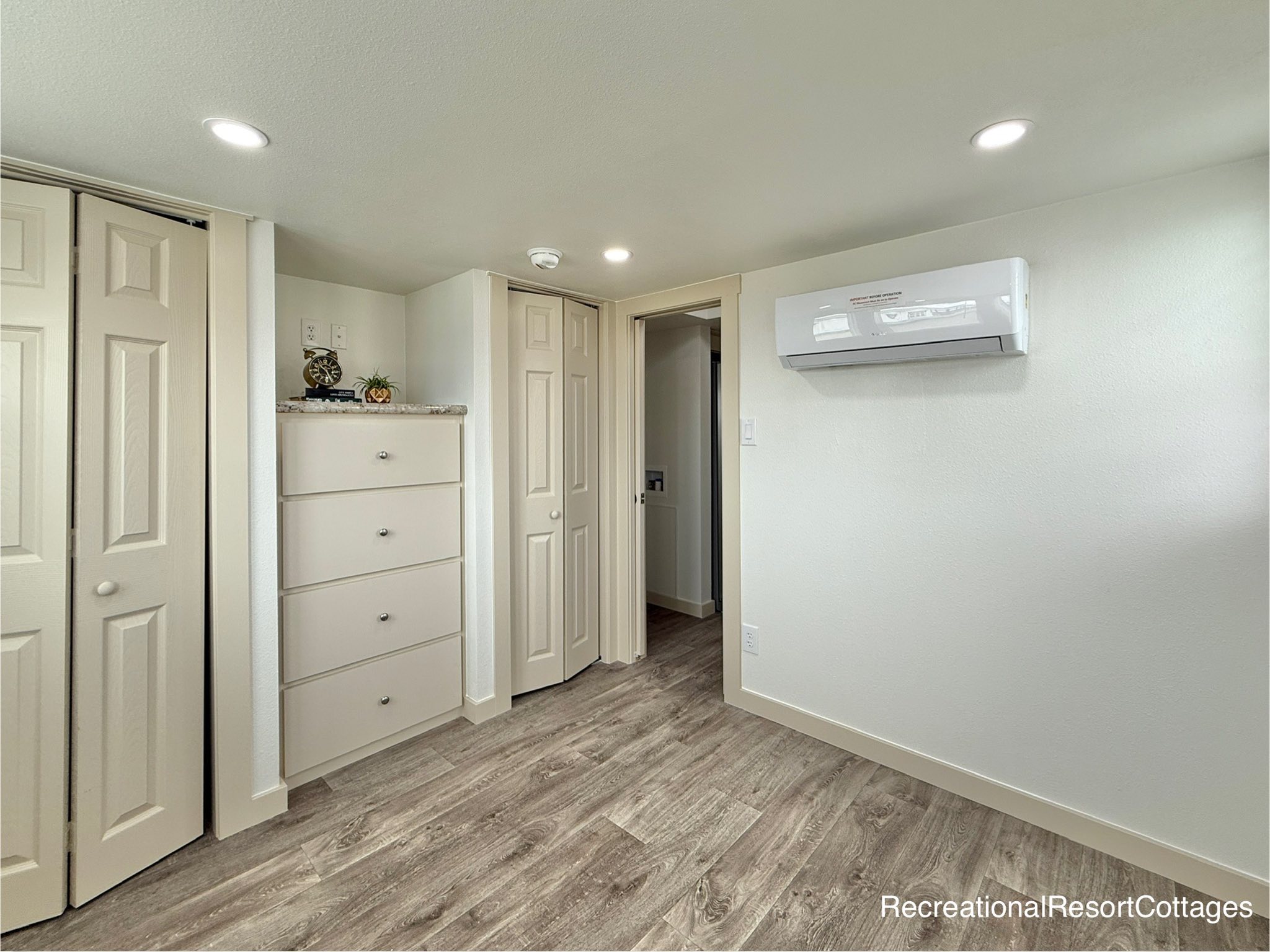 A small, minimalist room features light-colored walls, wood flooring, and recessed lighting. A dresser and folded closet door are on the left, with a wall-mounted air unit on the right.
