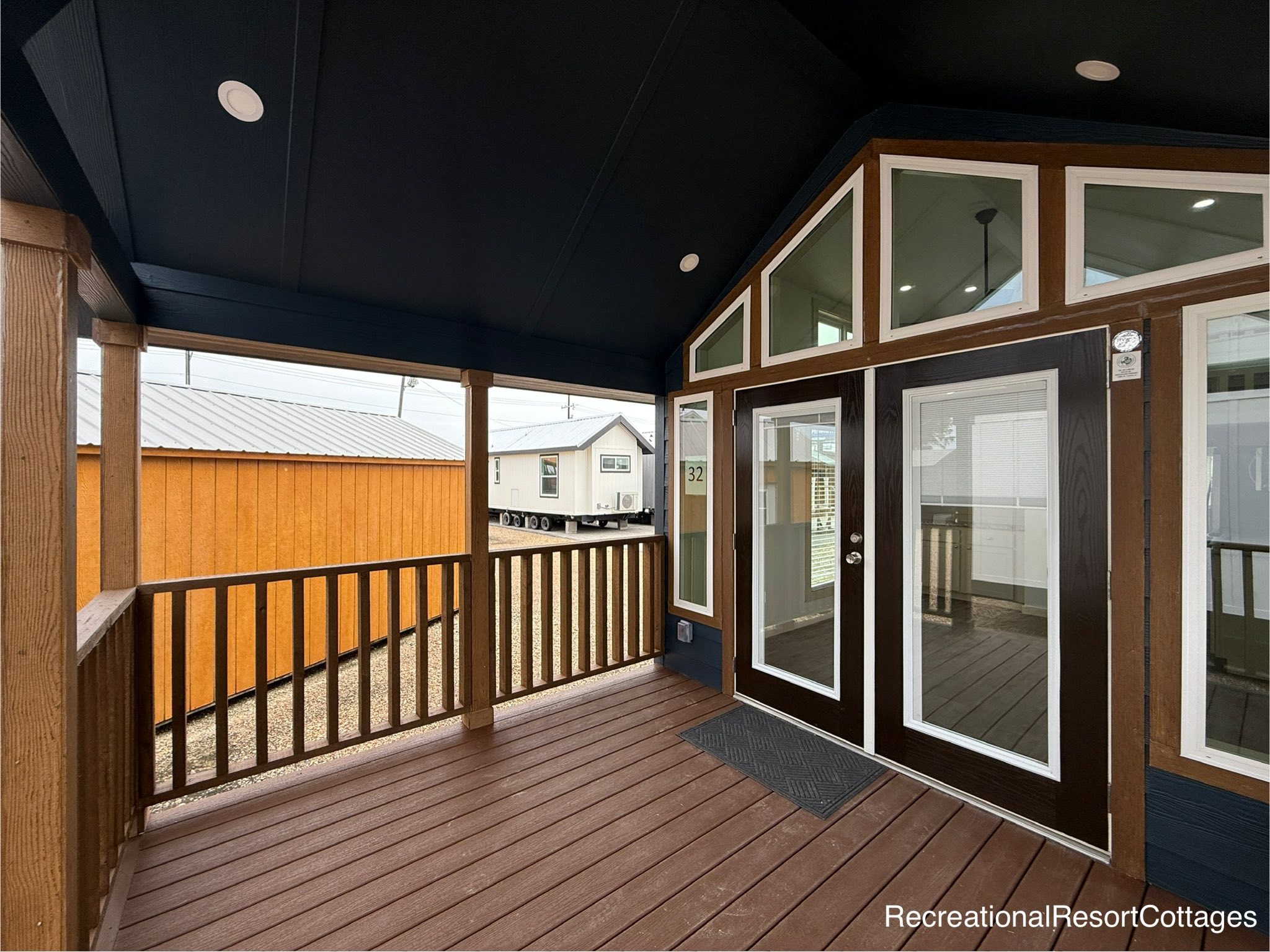 Covered porch with wood railing and brown decking attached to a small cabin with large windows and French doors, conveying a cozy, inviting feel.