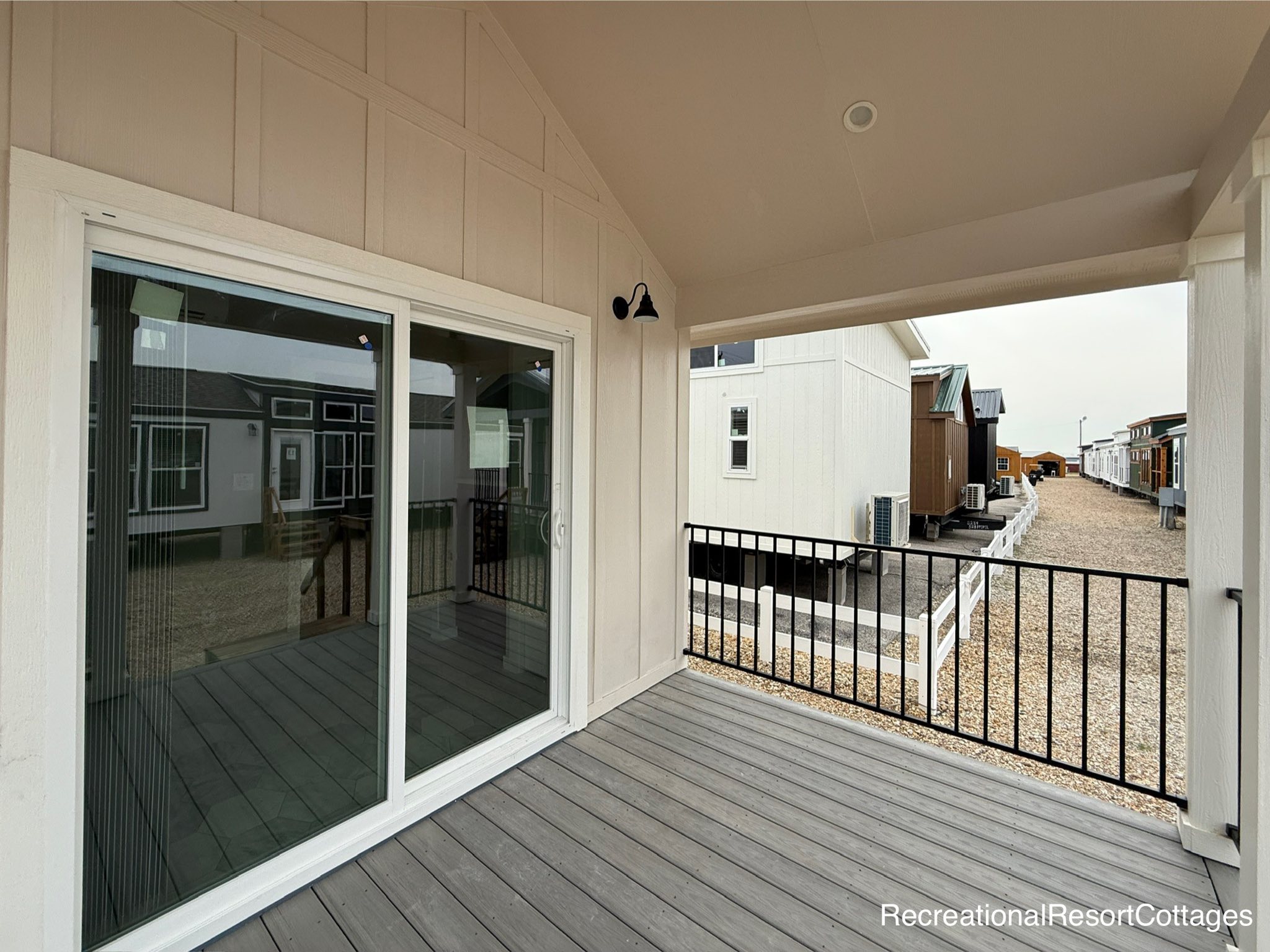 Covered porch with gray decking features a sliding glass door, black wall sconce, and iron railing. Views of tiny homes with gravel paths. Calm ambiance.