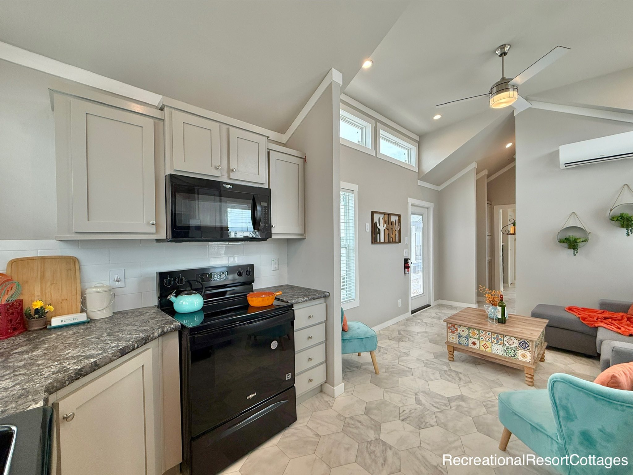 Bright and modern tiny home interior with a compact kitchen featuring light gray cabinets, black appliances, and granite countertops. The living area has vibrant turquoise chairs, a patterned coffee table, and gray sofa, creating a cozy and inviting atmosphere.
