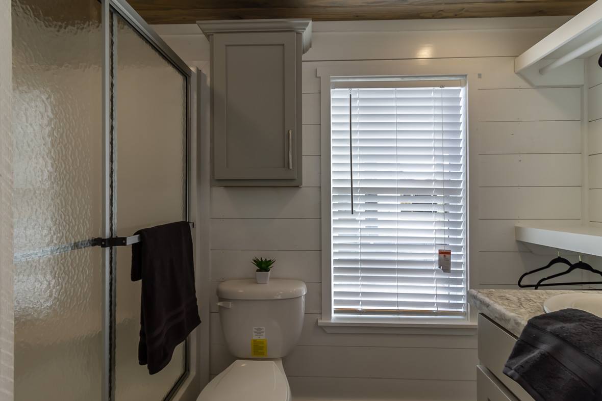 Small bathroom with a shower, white walls, and a window with blinds. A plant sits on the toilet tank, next to a dark towel on a rail. Calm, minimal vibe.