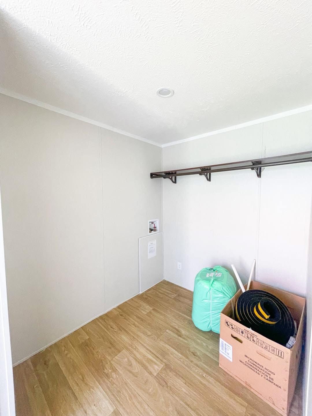 Small empty closet with light wood flooring and white walls. A wooden shelf runs along one wall. A box and green bag are on the floor, suggesting storage.