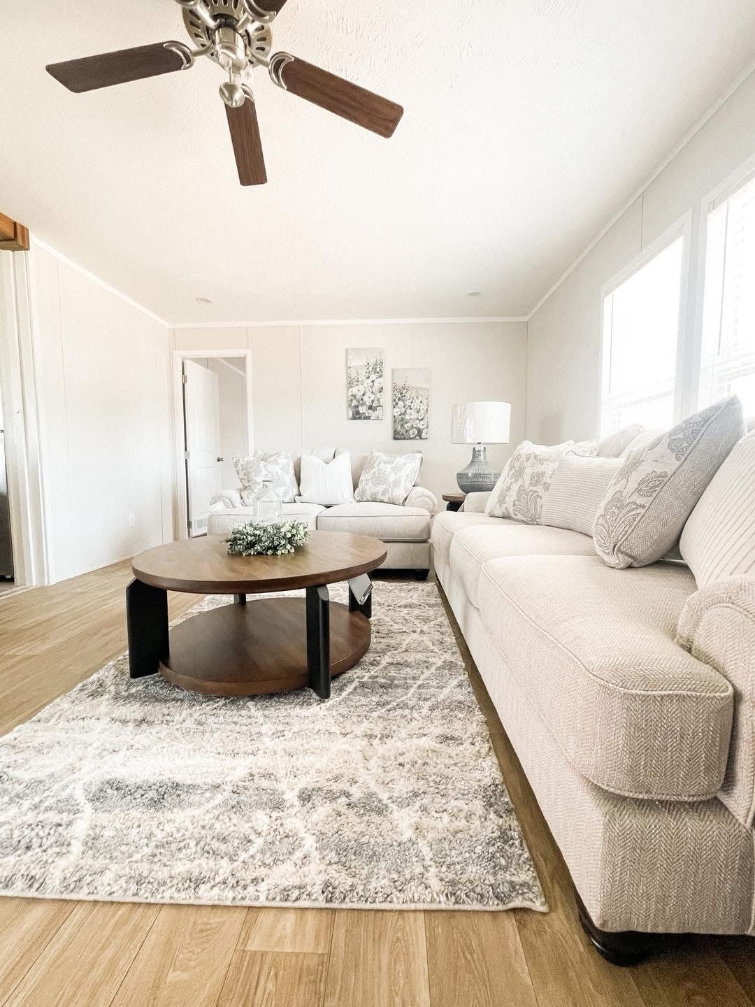 A bright, cozy living room with a beige sofa and armchair, a wooden coffee table on a patterned rug, wall art, table lamp, and a ceiling fan above.