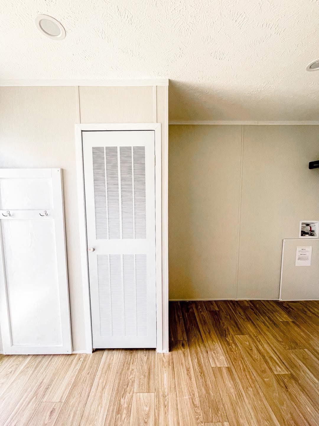 A bright room with light wood flooring and beige walls. A white louvered door and wall hooks are on the left, evoking a minimalist, clean ambiance.