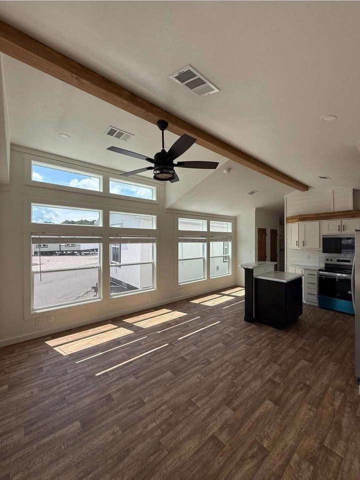Spacious interior of a modern kitchen and living area with tall windows, wooden flooring, and a ceiling fan. Sunlight streams in, creating a warm ambiance.
