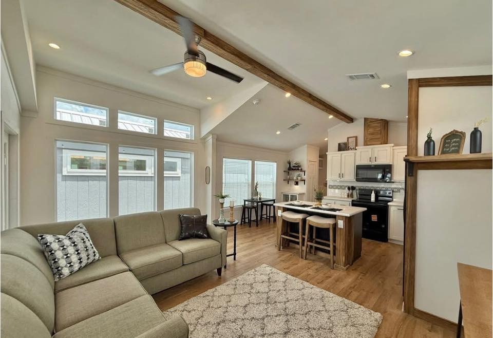 Spacious, modern open-plan living room and kitchen. Cozy L-shaped sofa, wooden beams, ceiling fan, bright with large windows, neutral tones.
