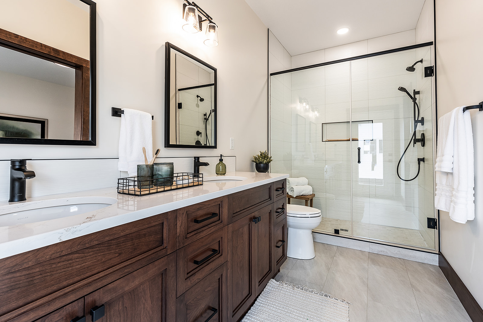 Modern bathroom with dark wood vanity, marble countertop, dual mirrors, and black fixtures. Glass-enclosed shower and potted plant add elegance.