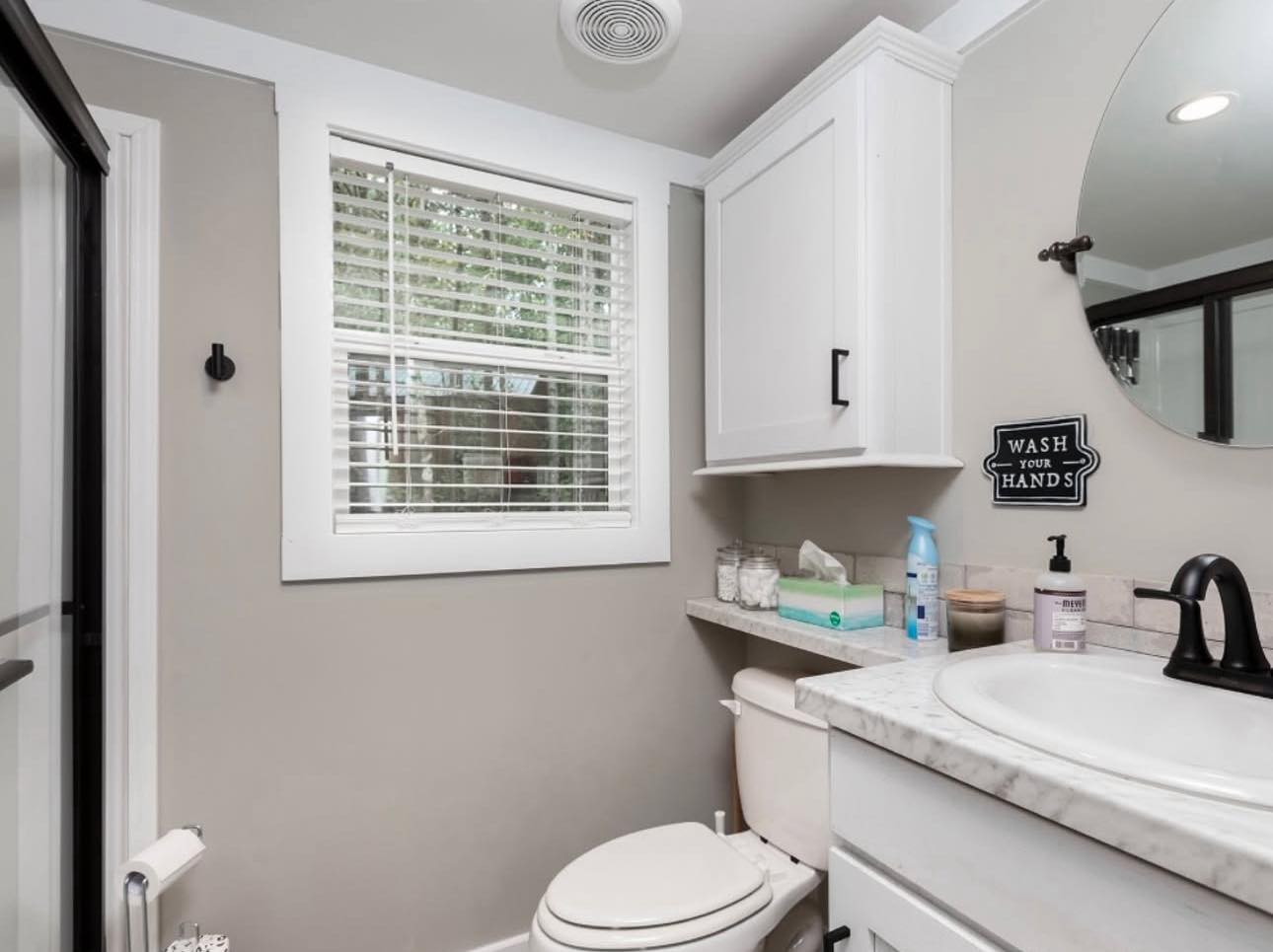 A small, modern bathroom with beige walls and a white window. Features a toilet, marble countertop with toiletries, round mirror, and a "Wash Your Hands" sign.