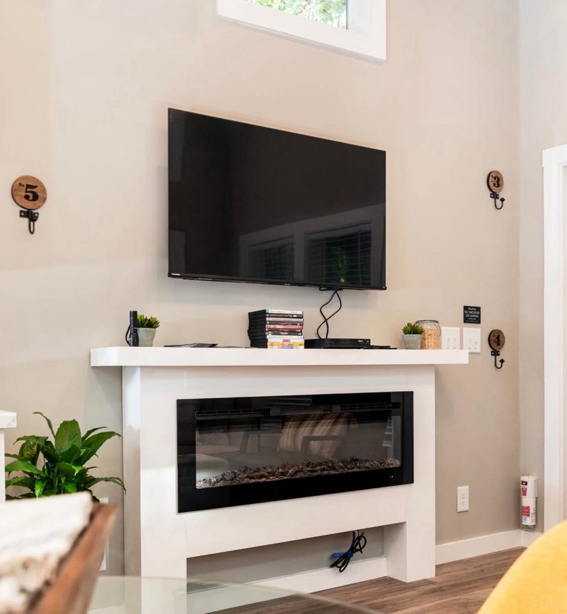 A modern living room corner with a mounted flat-screen TV above a white mantel and electric fireplace. Potted plants and a stack of games add a cozy, tech-savvy vibe.