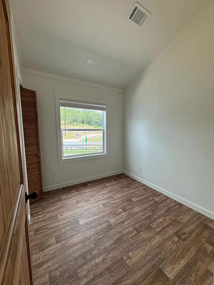 An empty room with white walls, wood flooring, a small window showing a grassy area outside. A wooden door is ajar, creating a calm atmosphere.