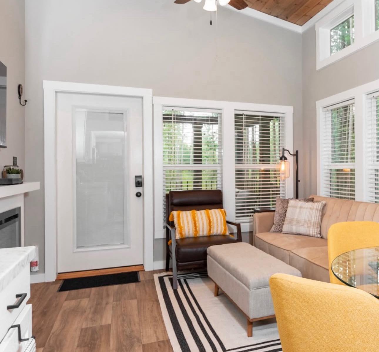 Cozy living room with a light beige sectional sofa, yellow and gray pillows, and a modern brown chair. Large windows bring in natural light.