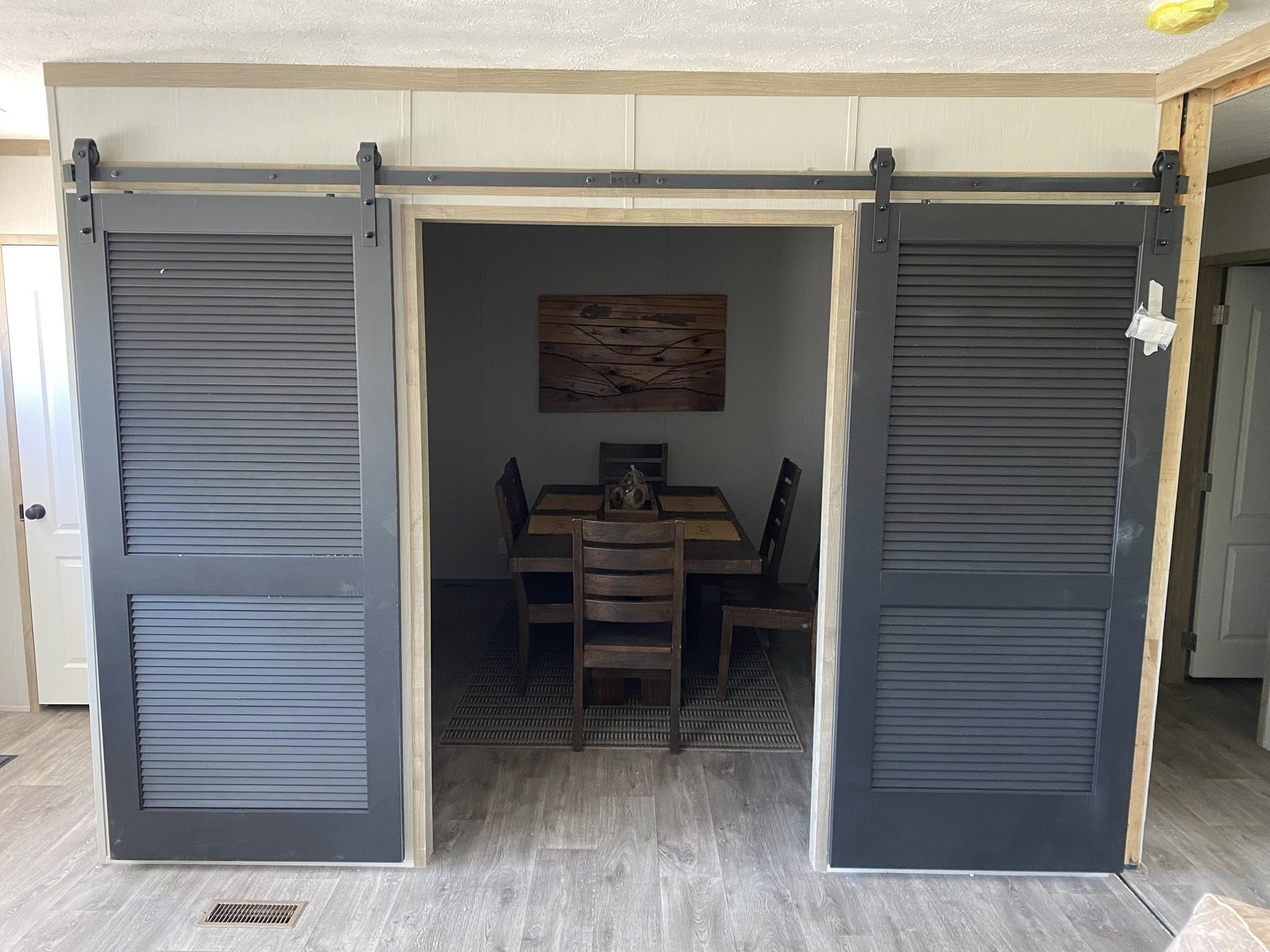 Dark gray sliding barn doors open to reveal a cozy dining room with a wooden table, chairs, and a decorative wall piece, creating a rustic, welcoming atmosphere.