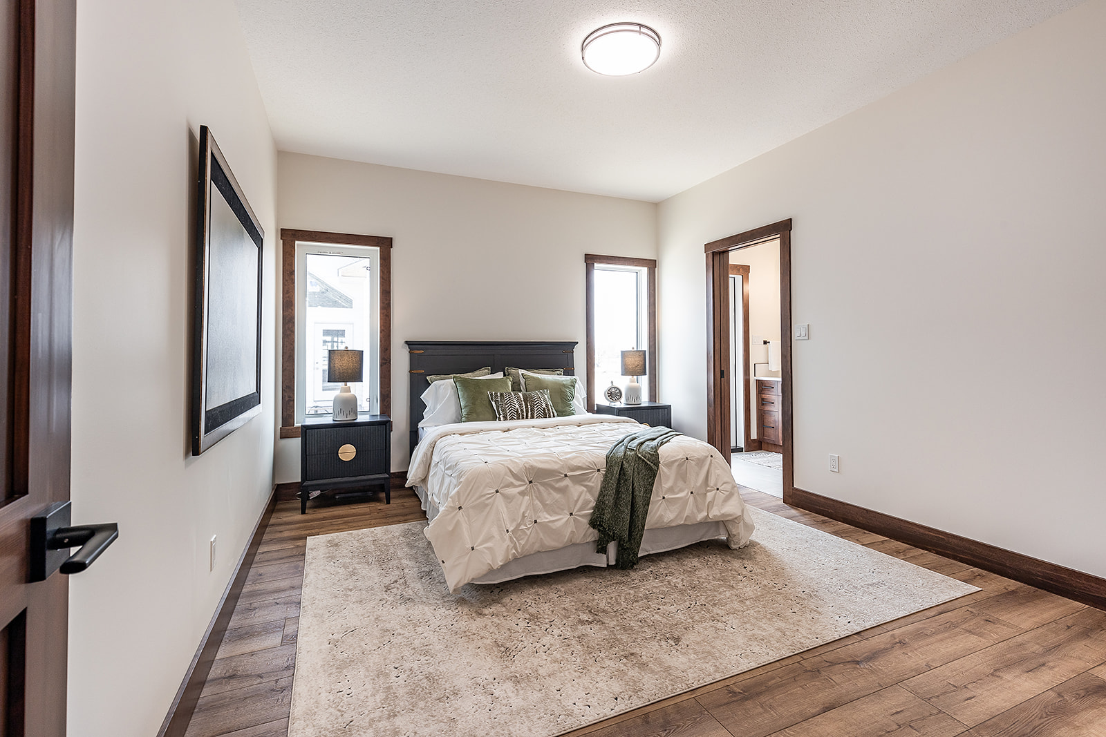 Modern bedroom with wooden floor, white walls, and a central bed adorned with green cushions and throw. Two windows flank the bed, adding light.