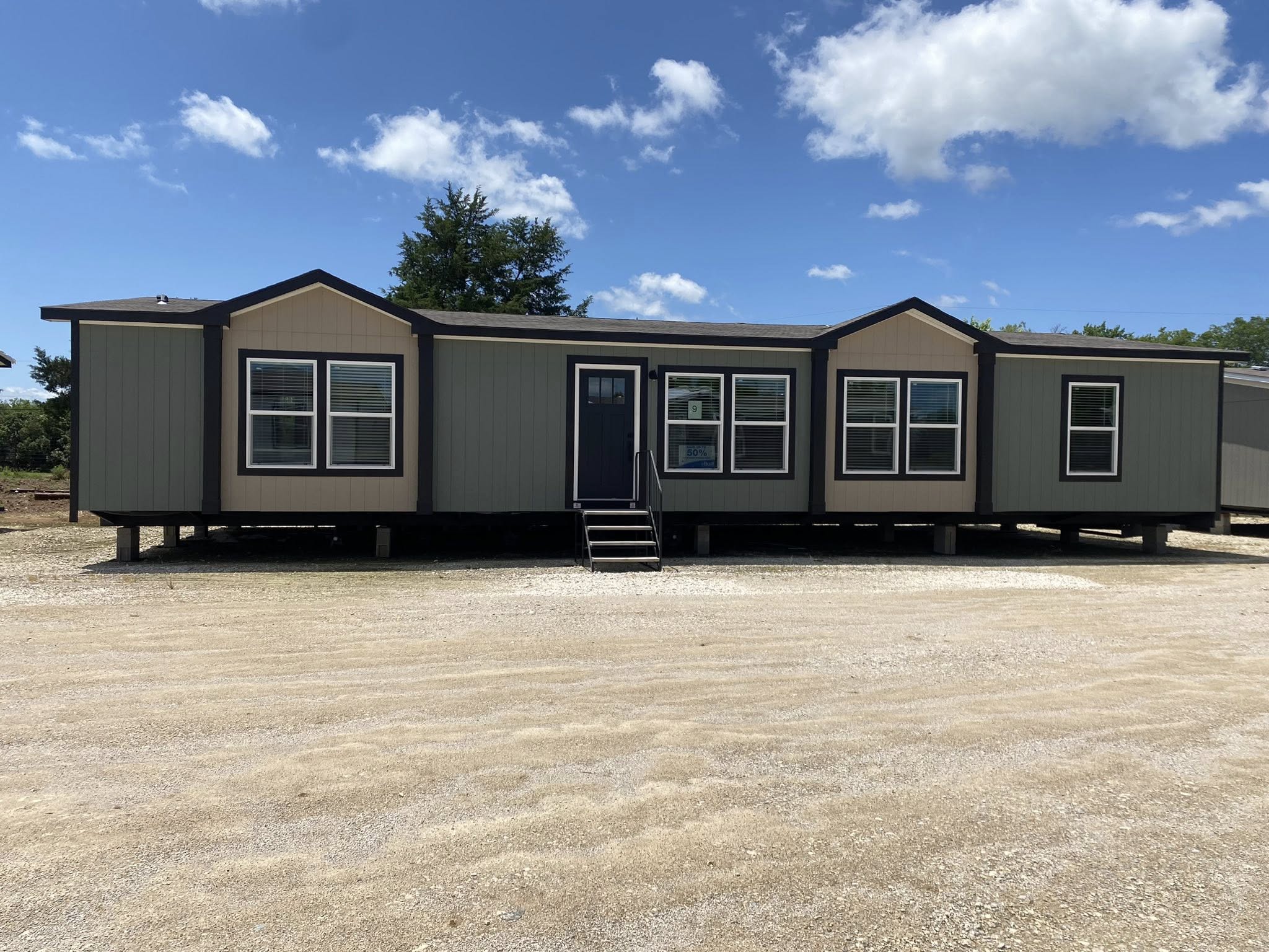 A large beige and gray mobile home with a black door and several windows sits on a gravel lot under a bright blue sky with scattered clouds.