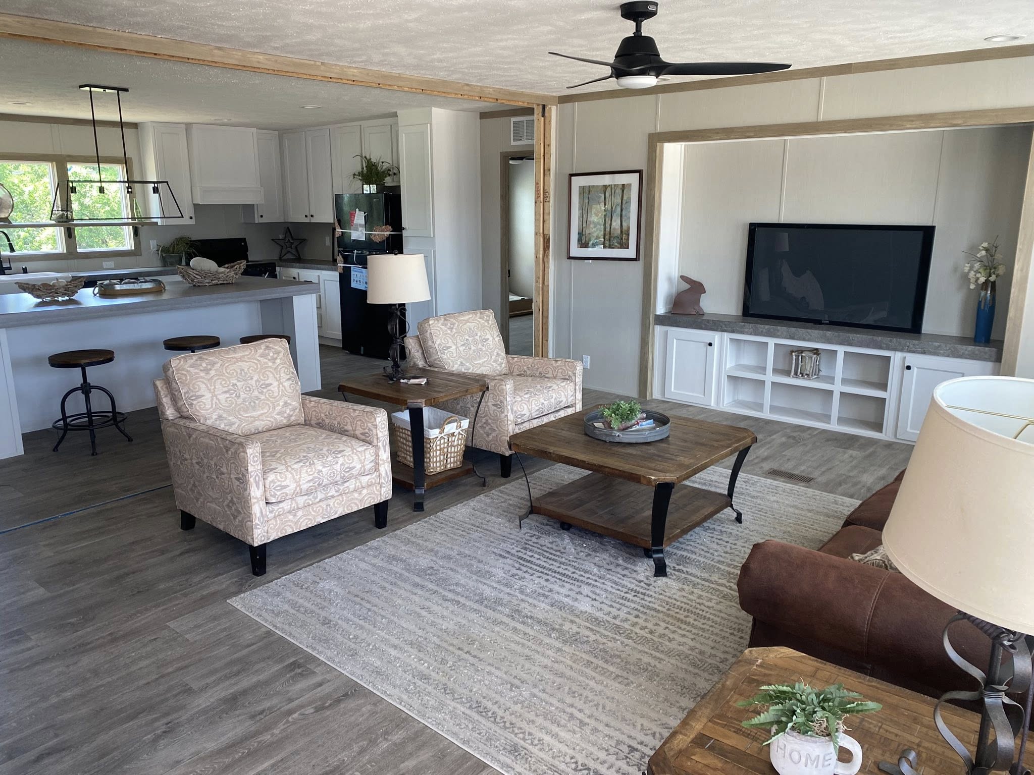 Modern living room with neutral tones. Two patterned armchairs face a wooden coffee table, adjacent to a sofa. Open kitchen with bar stools in the background.