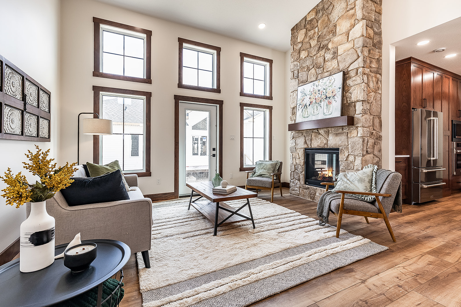 Cozy living room with tall windows, stone fireplace, and modern decor. Neutral tones, soft rug, and plants create a welcoming, serene ambiance.