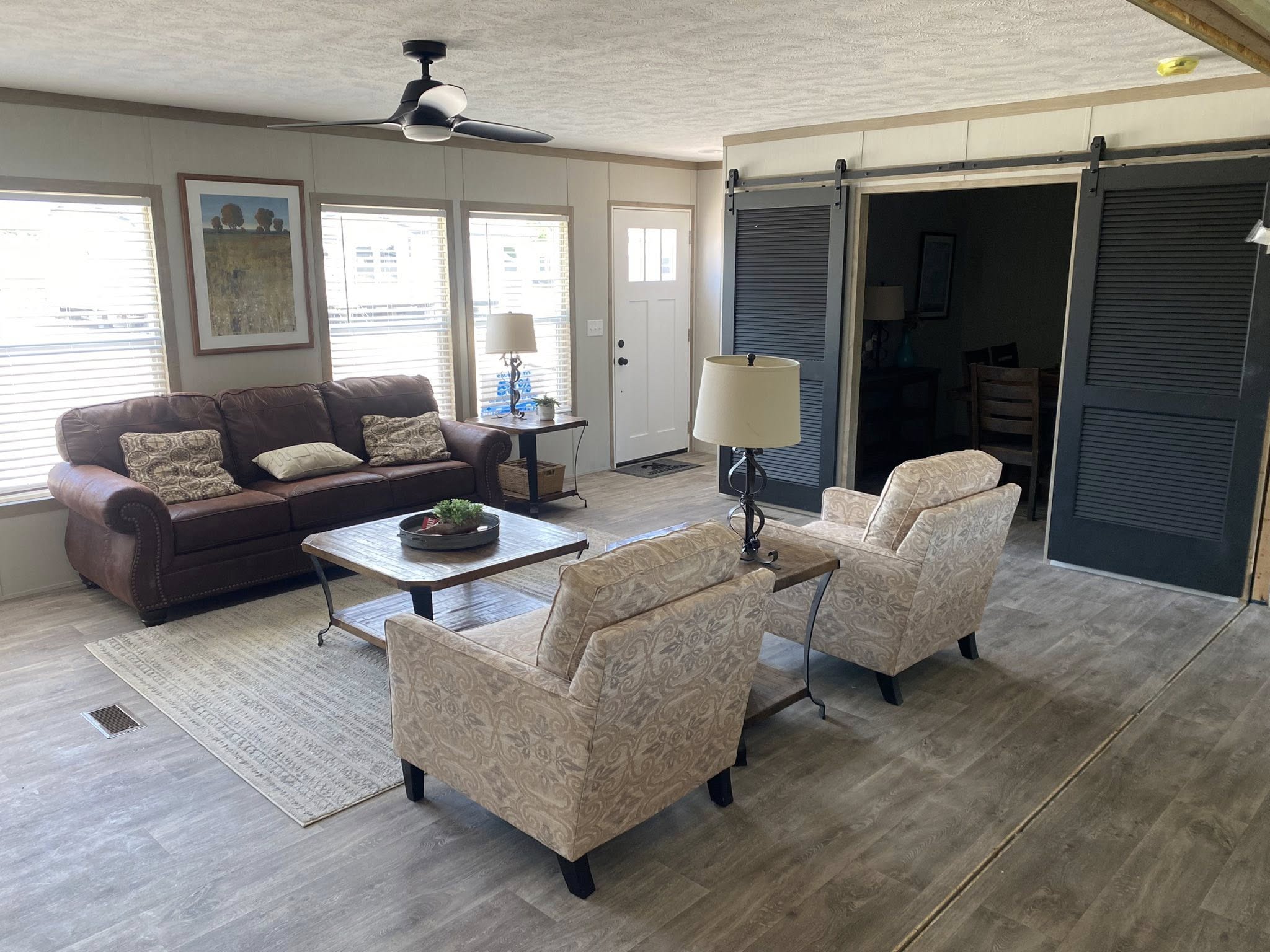 Cozy living room with a brown leather sofa, patterned armchairs, and a wooden coffee table on a beige rug. TV on a white console; framed art decor.