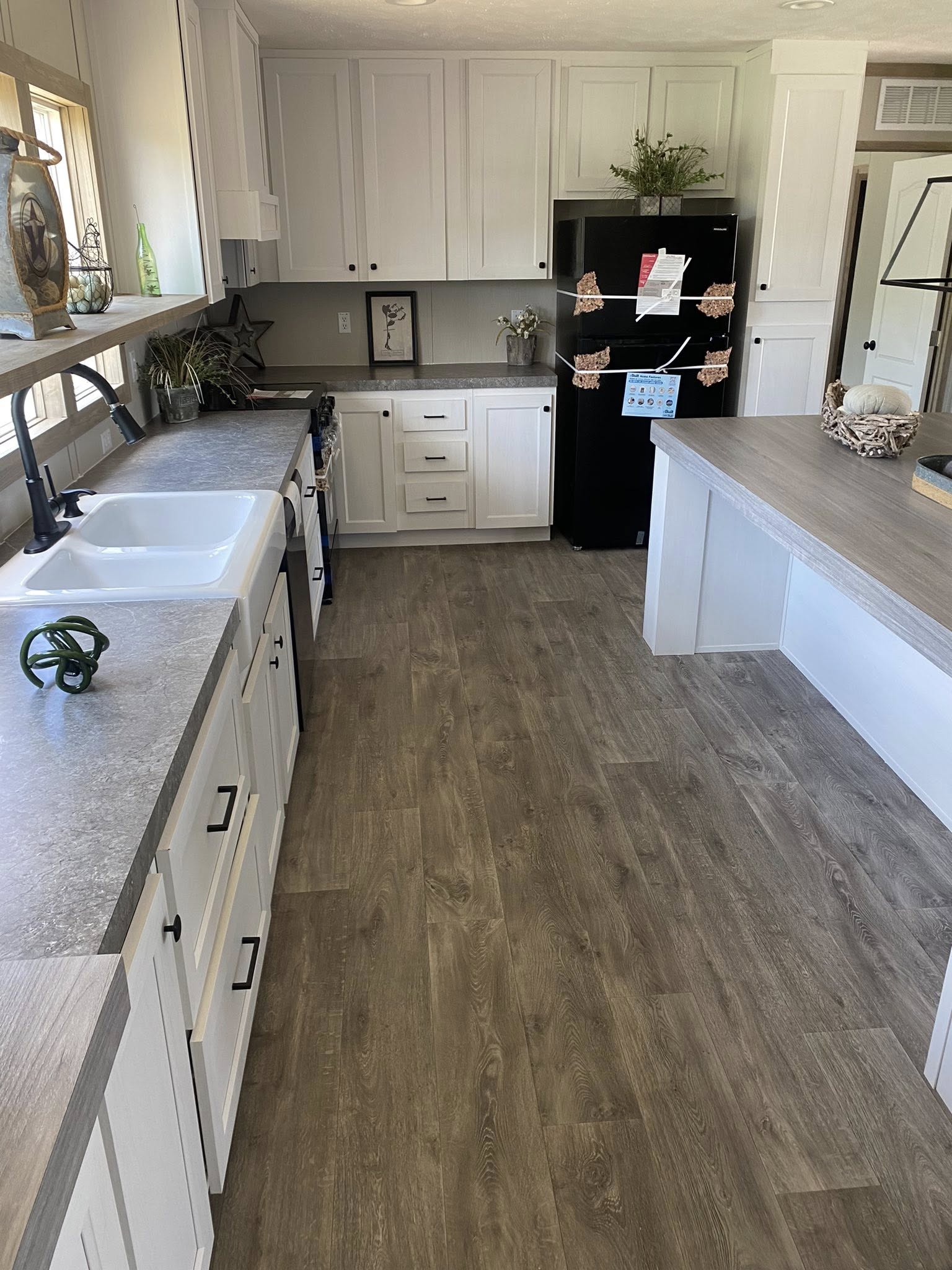 Stylish kitchen with light wood flooring, white cabinets, and grey countertops. A black fridge, decorative plants, and natural light create a welcoming space.